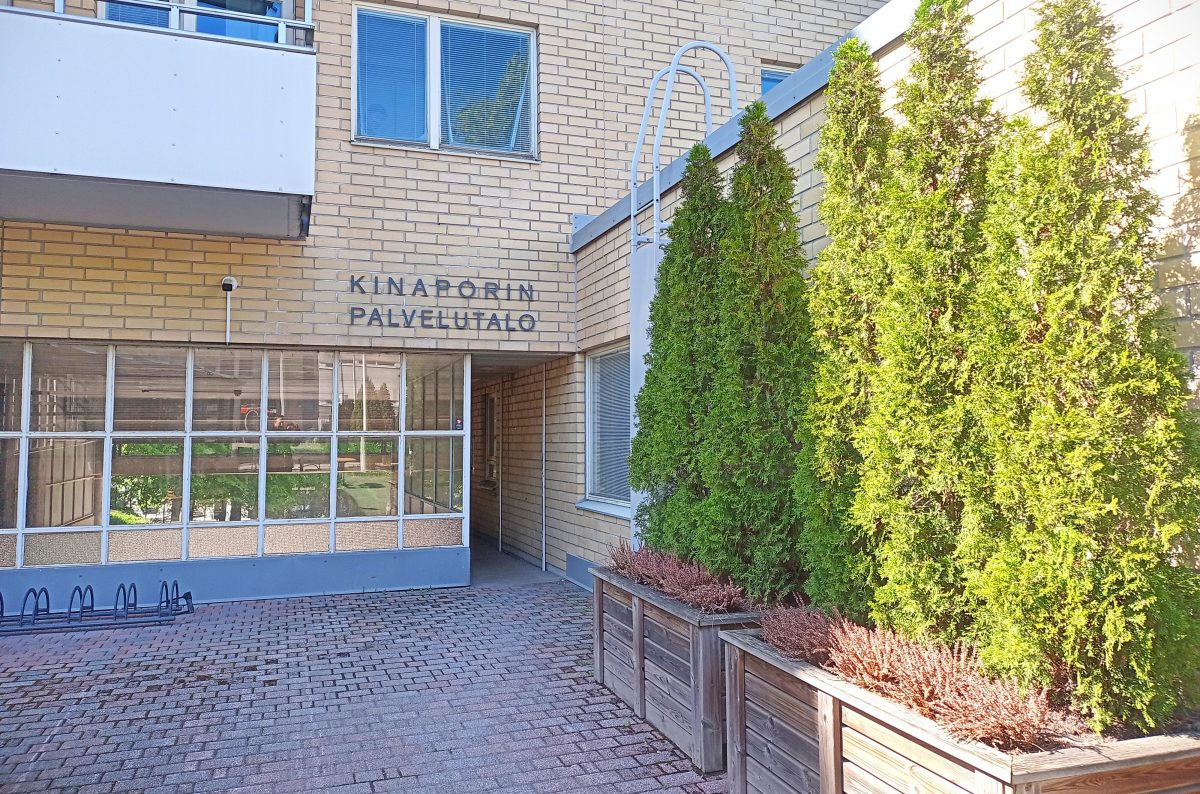 Kuvassa näkyy Kahvila Aleksiksen ikkunoita. Yläpuolella lukee "Kinaporin palvelutalo". Etualalla on vihreää kasvillisuutta.