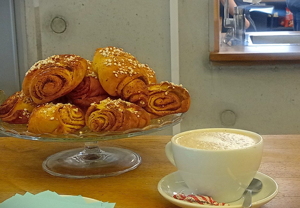 Kuvassa on Månsas Deli -kahvilan palvelutiski, jossa edustalla capuccino-kuppi ja taustalla jalallinen lasitarjoitin täynnä korvapuusteja.